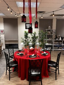 An elegantly set dining display in a showroom, featuring a round table with a rich red tablecloth and black chairs with a lattice back design. Red fabric ribbons are draped overhead, with black lanterns adding to the decor. The table is set for four, with black plates, red napkins folded into a hat shape, and gold-rimmed stemware. Green bamboo plants flank the table, enhancing the Asian-inspired aesthetic of the setup.