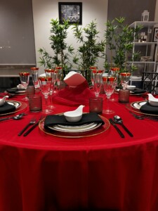 Close-up view of a festive dining table setup showcasing a vibrant red tablecloth. Each place setting includes elegant black and gold-rimmed plates, black napkins, and unique white napkin rings shaped like a bird. The table features gold-rimmed glassware and red votive candle holders that cast a warm glow. Lush green bamboo plants in the background complement the Asian-inspired theme,