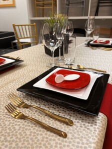 A sophisticated table setting for a Lunar New Year celebration, with a layered place setting consisting of a black square charger plate, a smaller white square plate, and a vibrant red round plate on top, accented with a whimsical white bird-shaped napkin ring. In the foreground, two gold forks complement the setup, with a sparkling glass water cup, wine glass, and clear vase with greenery adding elegance. The tablecloth beneath has a subtle gold-flecked pattern, adding a festive sparkle to the occasion